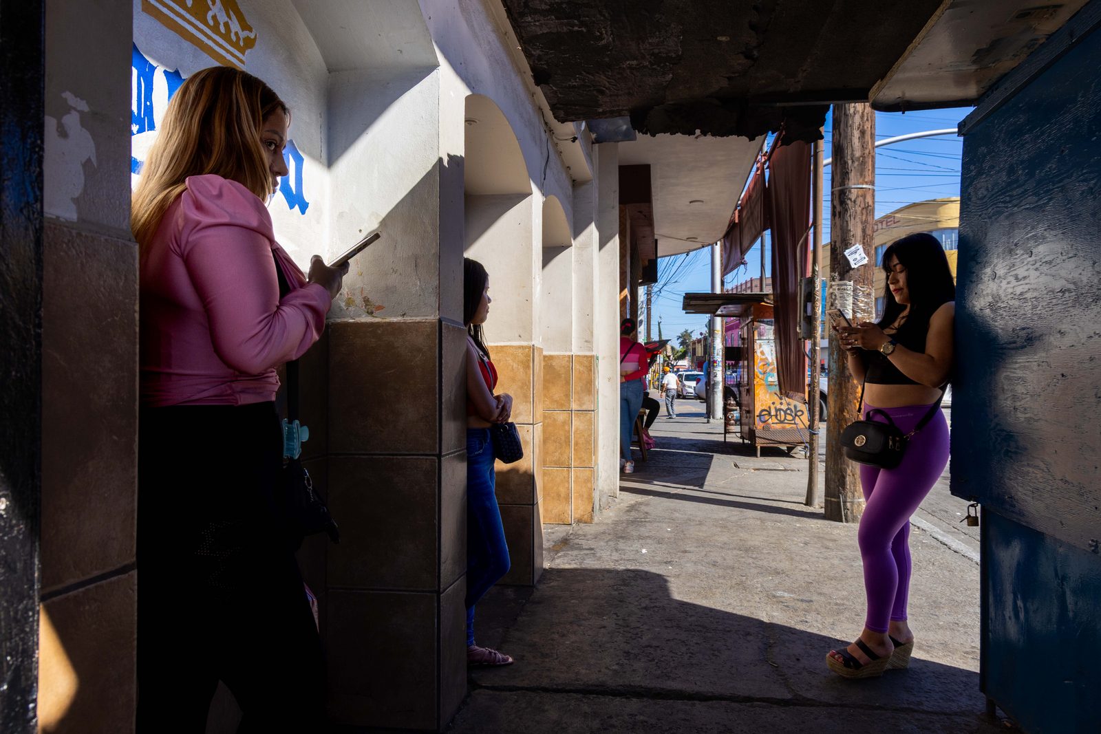 A covered walkway in hard sunlight, a woman in pink standing at the left, figures gathered in the distance — The Liminal Innocence