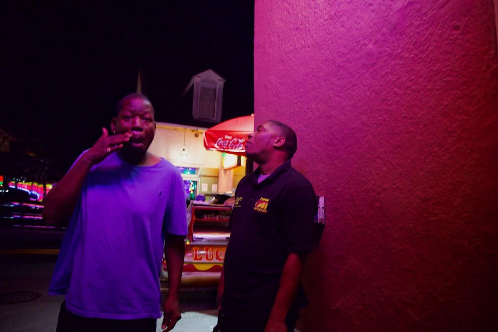 Two men on a night street beside a magenta-lit wall, a food cart glowing behind them — New Orleans