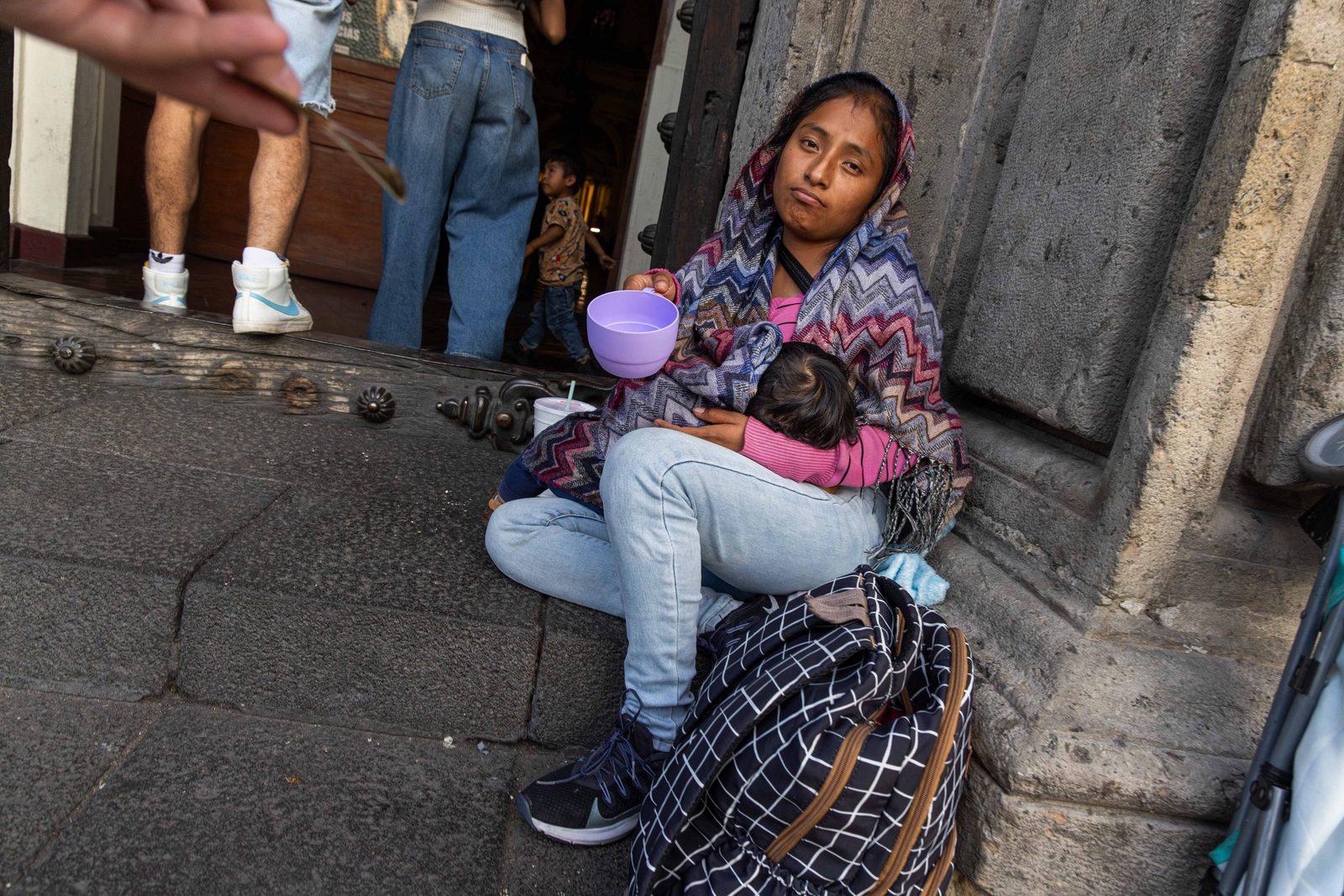 A woman sits on stone steps holding a baby, a purple bowl in her hand, a patterned bag beside her — The Liminal Innocence